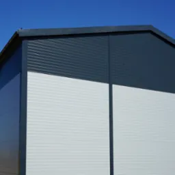 Zewnętrzny widok rogu nowo wybudowanej hali magazynowej z szarej blachy trapezowej i białych płyt warstwowych, widoczne rynny i błękitne niebo w tle.