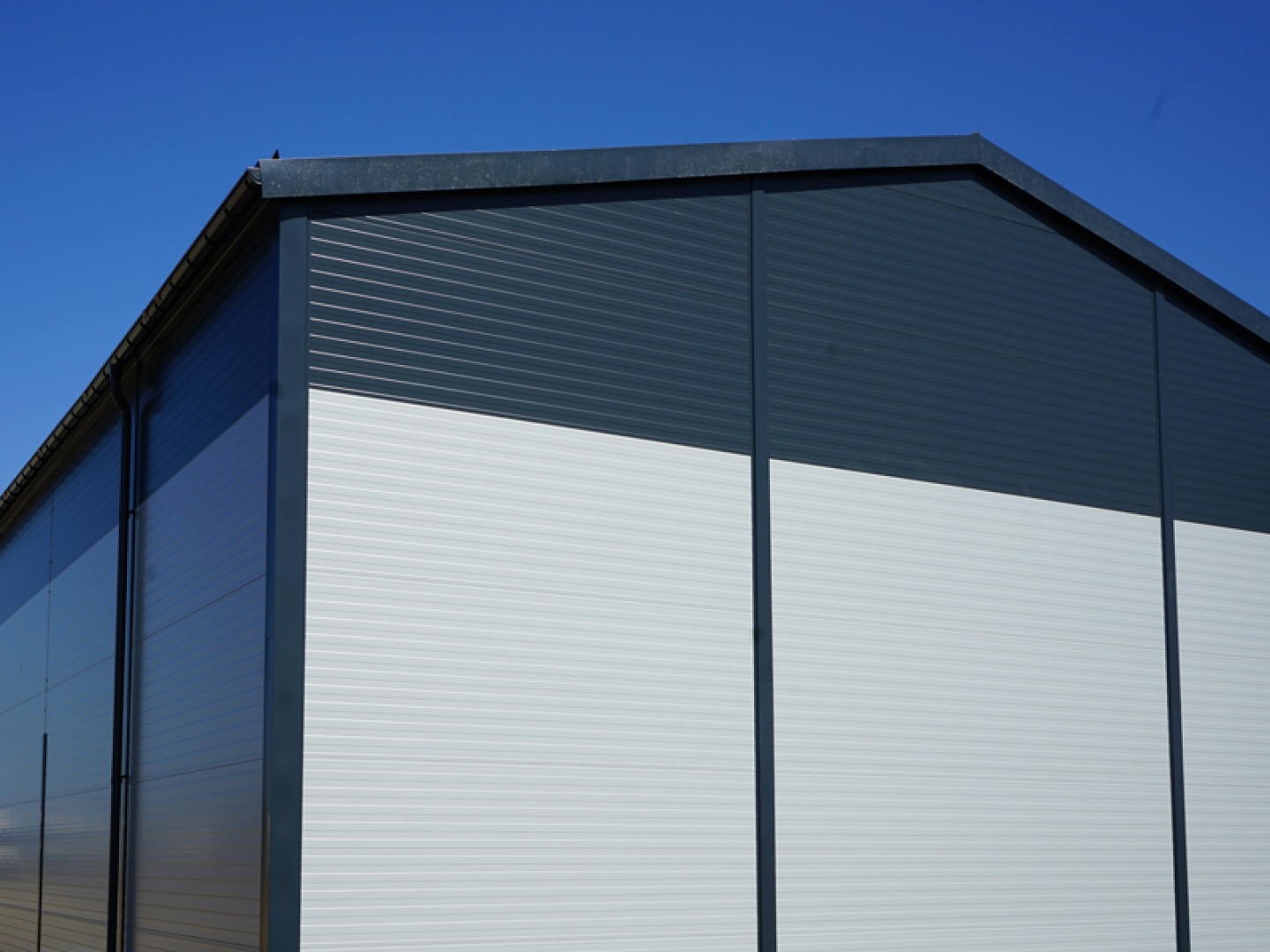 Zewnętrzny widok rogu nowo wybudowanej hali magazynowej z szarej blachy trapezowej i białych płyt warstwowych, widoczne rynny i błękitne niebo w tle.