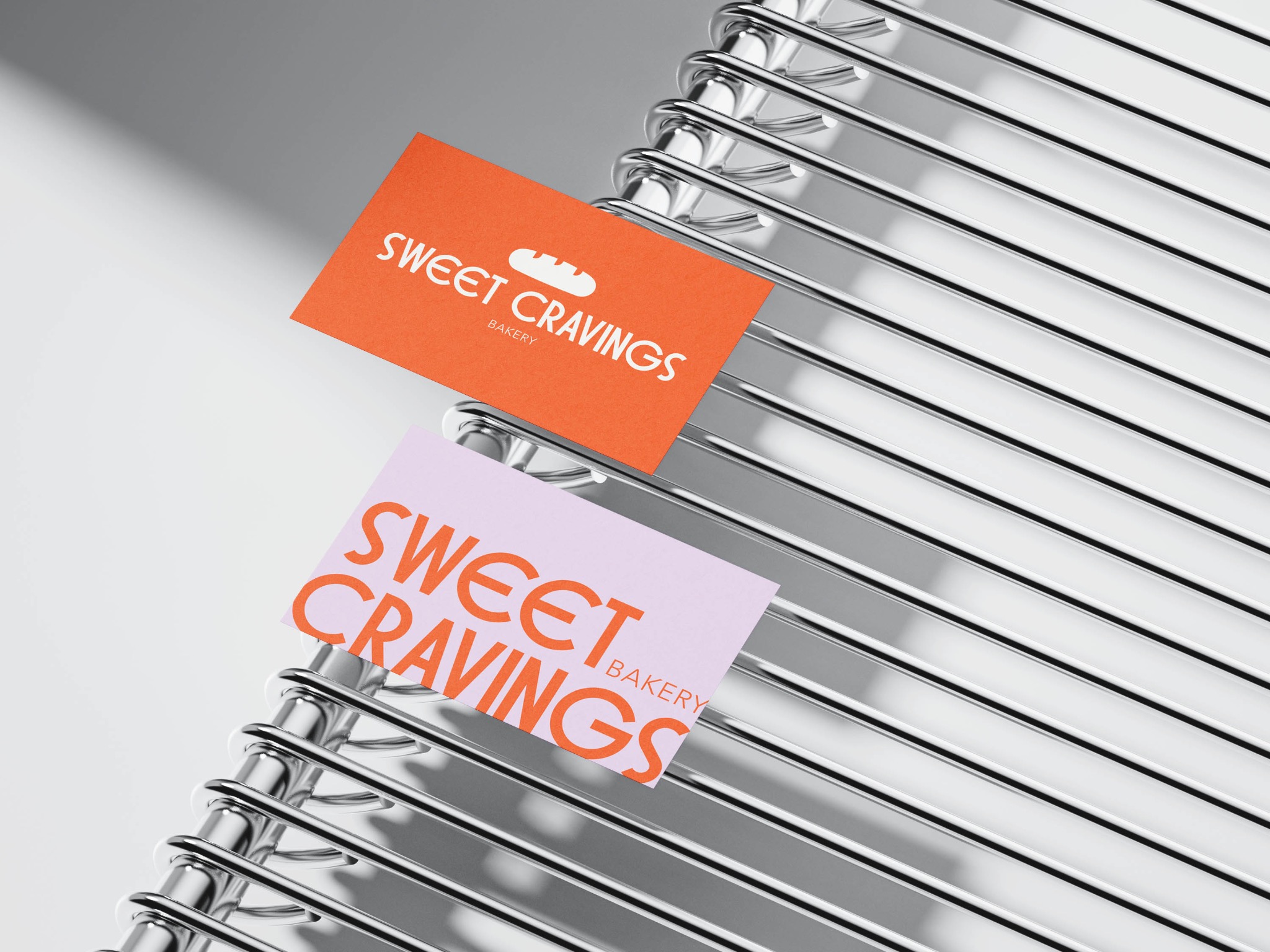 Dwie wizytówki piekarni 'Sweet Cravings' w kolorach pomarańczowym i liliowym, ułożone na metalowej konstrukcji z prętów, z widocznym cieniem.