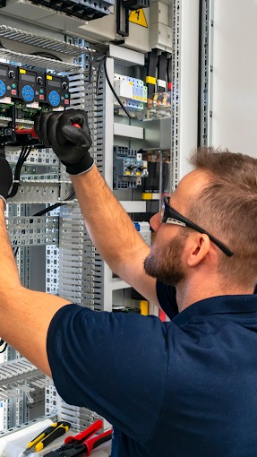 Elektryk w okularach, w Dongen, pracuje nad instalacją elektryczną w szafie sterowniczej, używając narzędzi leżących obok.