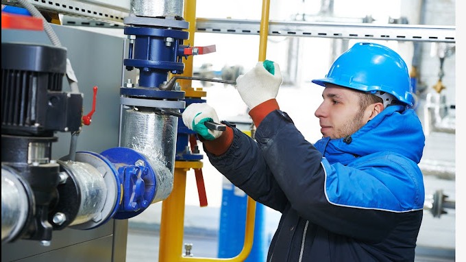 Pracownik w niebieskim kasku i kurtce dokręca śrubę na niebieskim zaworze rurociągu w instalacji przemysłowej, używając klucza nastawnego i rękawic roboczych.