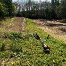 Maciej krypciak - Spalinowa kosa Stihl na trawie, w tle teren po niwelacji z widocznymi śladami opon i drzewami. Słoneczny dzień.