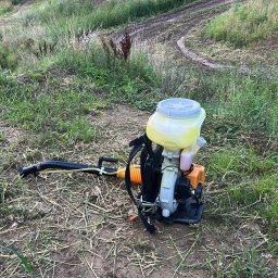 Maciej krypciak - Opryskiwacz spalinowy plecakowy leży na trawie na tle pagórkowatego terenu z drzewami. Sprzęt do pielęgnacji ogrodów.