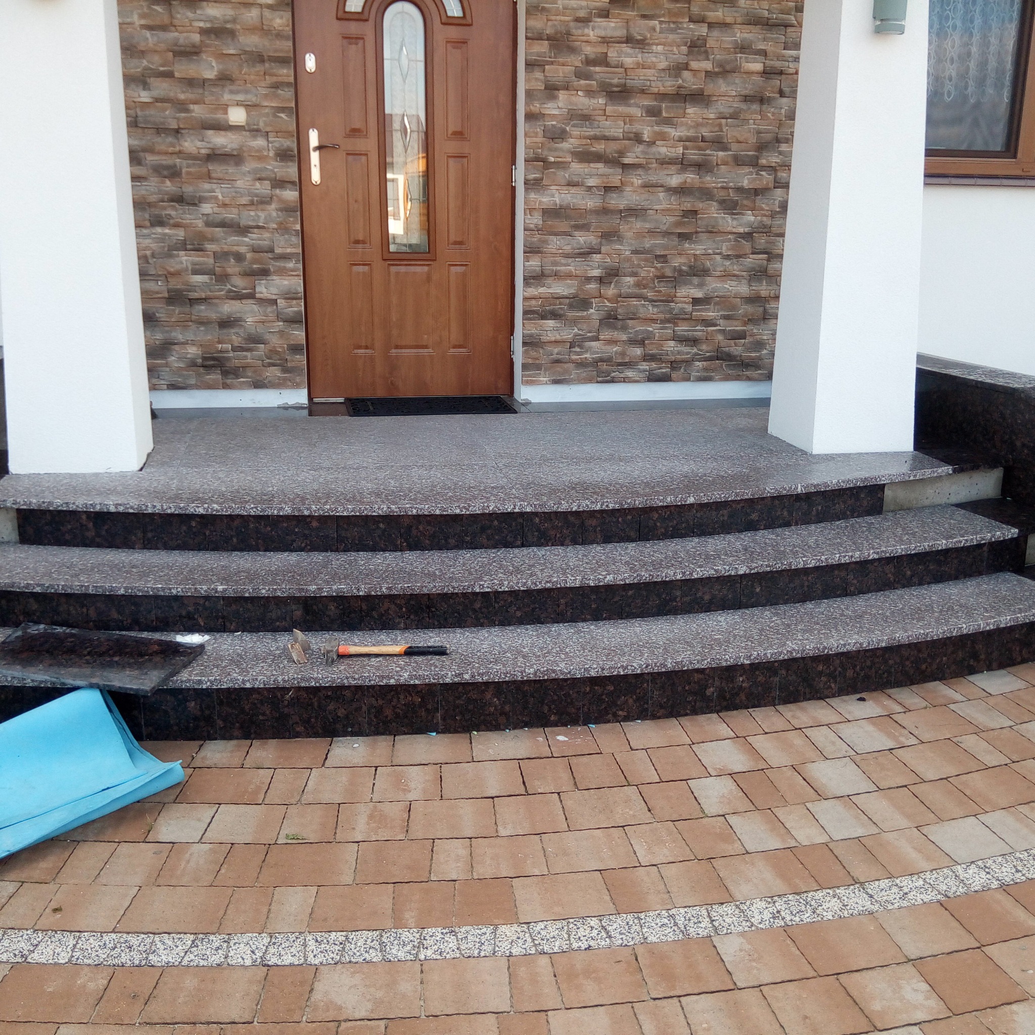 Wykończone schody wejściowe do domu z brązowego granitu, z widocznym młotkiem i niebieskim elementem, oraz elewacją imitującą kamień.