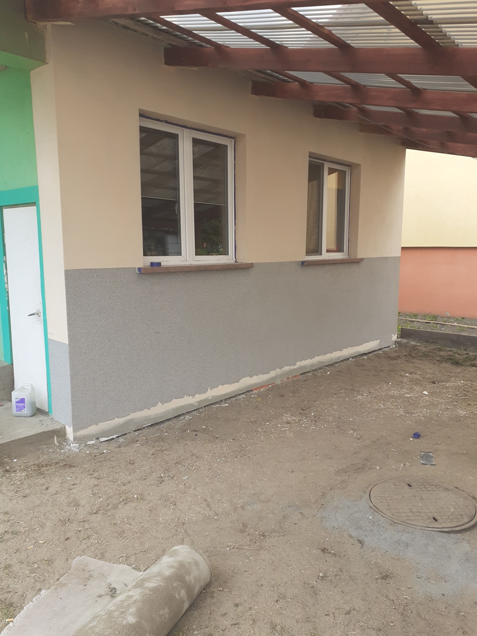 Fragment ściany budynku z oknami po nałożeniu dwukolorowej elewacji: górna część beżowa, dolna pokryta szarym tynkiem strukturalnym, widoczny fragment zadaszenia i teren wokół budynku w trakcie...