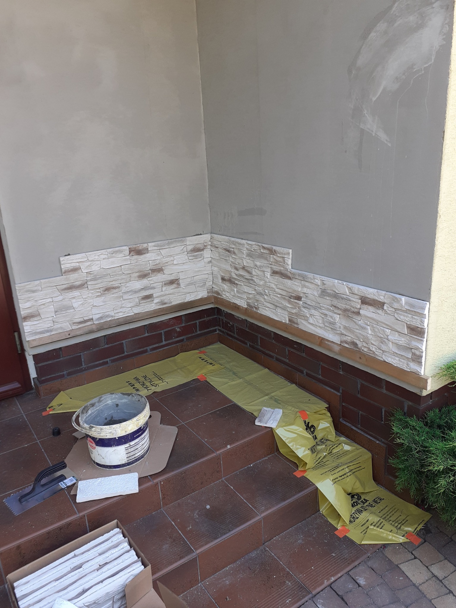 Fragment elewacji budynku w trakcie wykańczania imitacją kamienia, widoczne narzędzia, wiadro z klejem, karton z płytkami i folia ochronna na schodach.