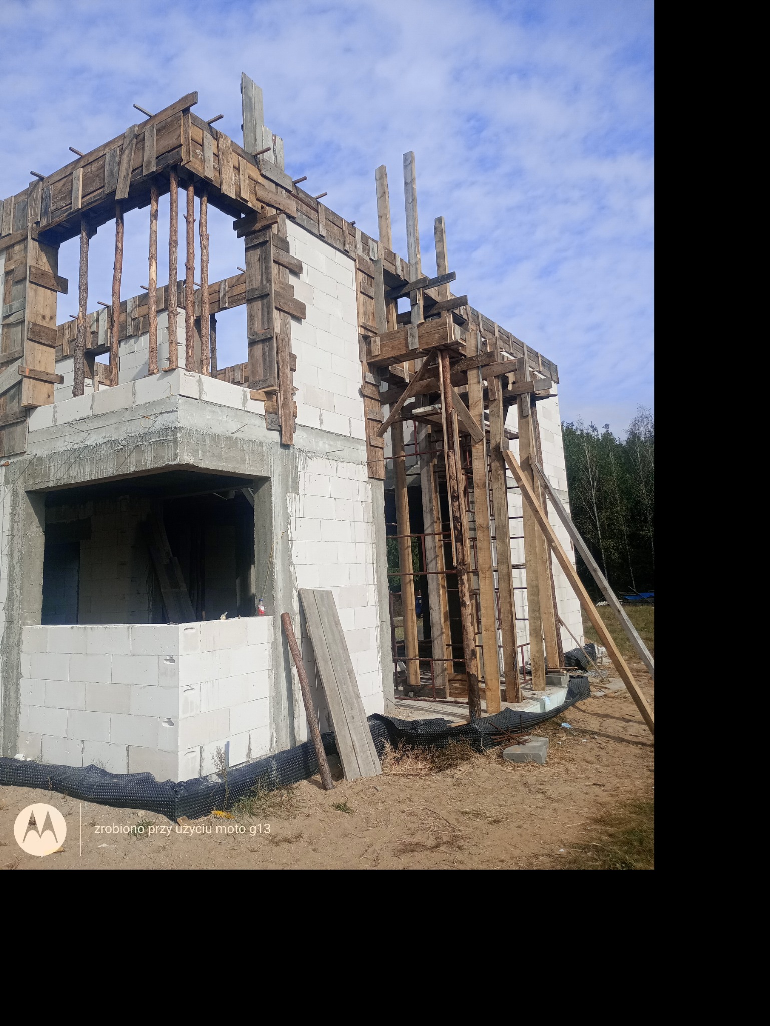 Surowy stan domu w trakcie budowy, ściany wzniesione z białych bloczków, drewniane szalunki, widoczne zbrojenia i deski, teren budowy.