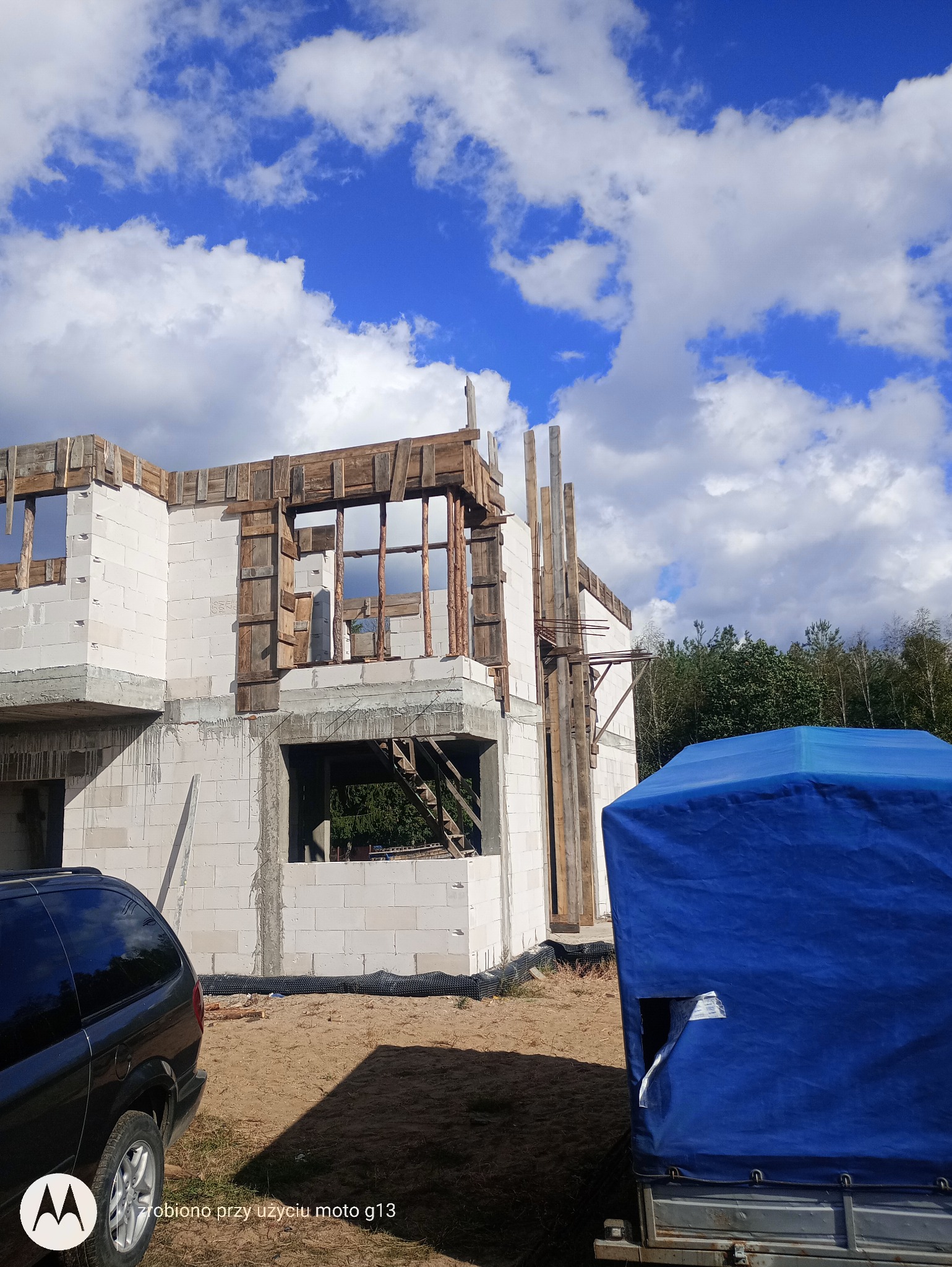 Wznoszenie dwukondygnacyjnego domu z białych bloczków, widoczne drewniane szalunki okienne i rusztowania, na pierwszym planie samochód i przyczepa z niebieską plandeką, w tle błękitne niebo...