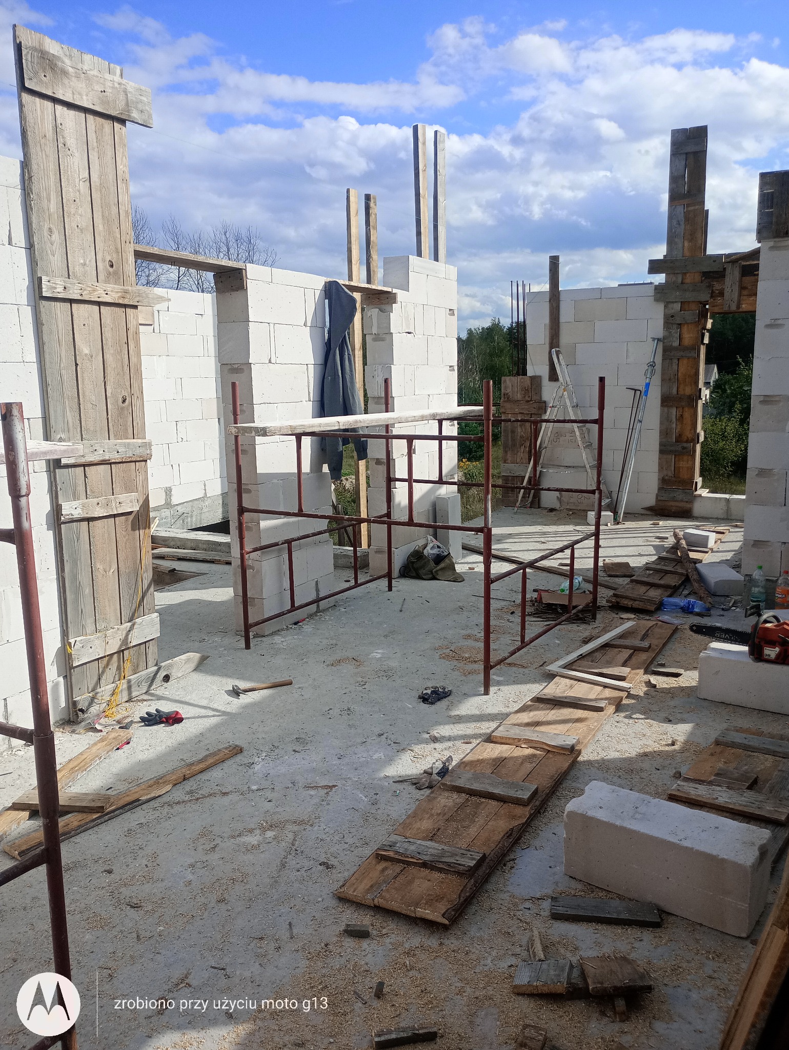 Wnętrze budowanego domu murowanego z białych bloczków, widoczne rusztowanie, drewniane elementy szalunkowe, narzędzia i resztki materiałów budowlanych na posadzce.