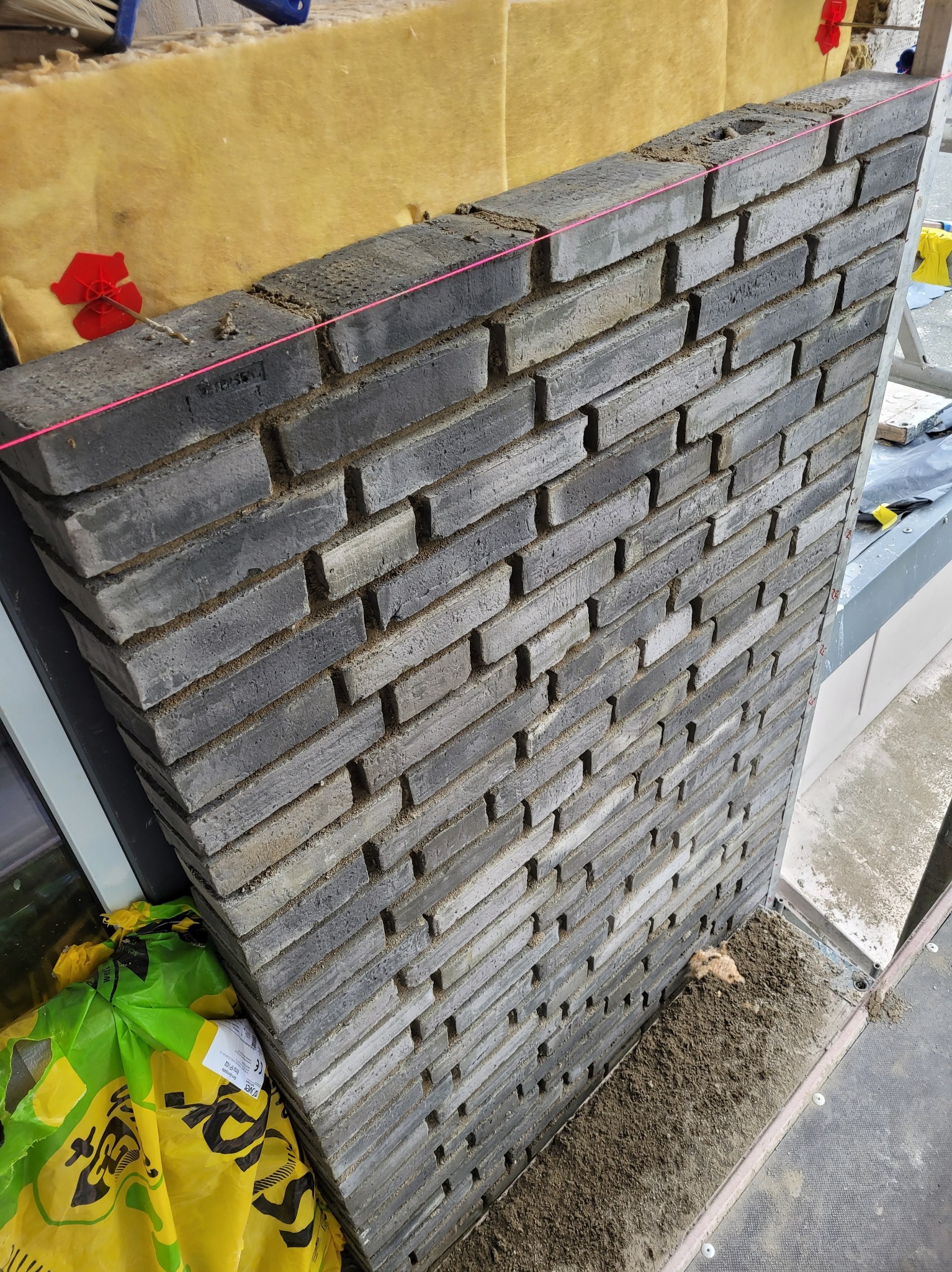 Wznoszona ściana z ciemnoszarych cegieł klinkierowych, widoczne spoiny i napięty sznurek murarski, ocieplenie z wełny mineralnej w tle, torba z materiałami budowlanymi u dołu.
