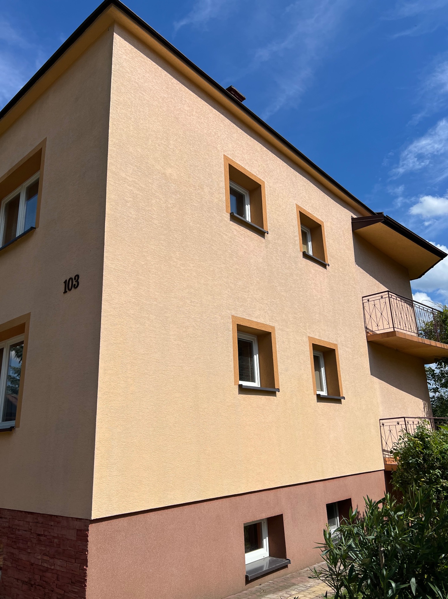 Dwupiętrowy budynek z beżową elewacją i brązowymi akcentami, numer '103' na rogu, balkony z metalowymi balustradami, widok z dołu w słoneczny dzień.