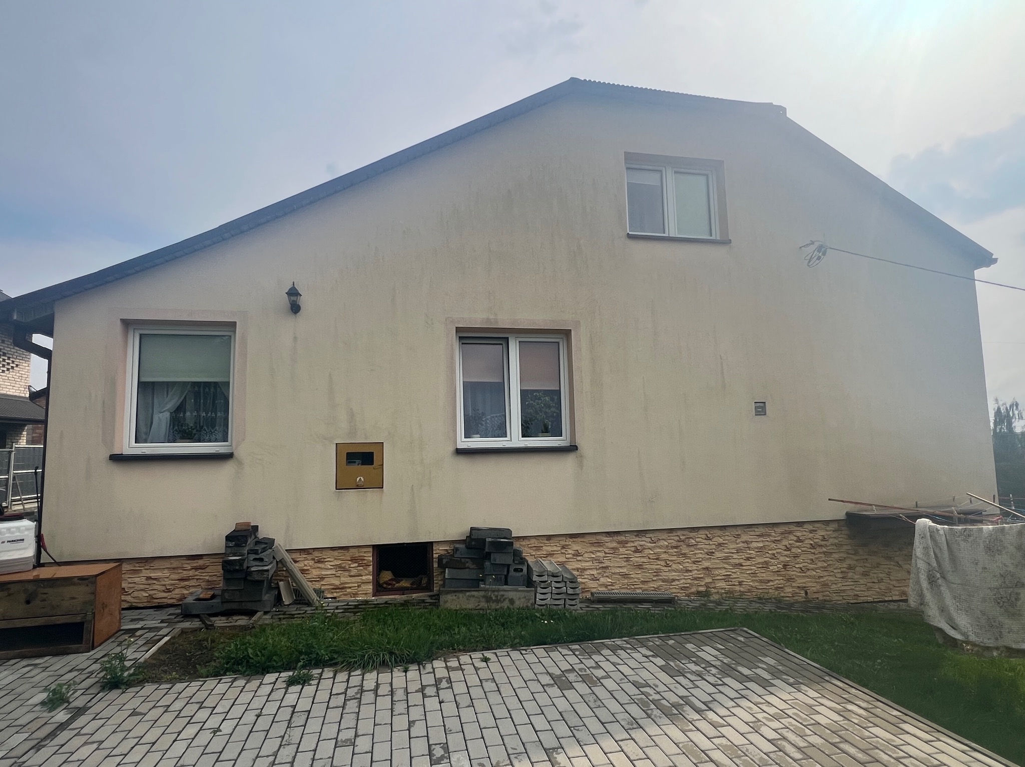 Dwukondygnacyjny dom z beżową elewacją, widoczne zabrudzenia i zacieki na ścianie, z kostką brukową przed budynkiem oraz stertą kostki przy fundamencie.