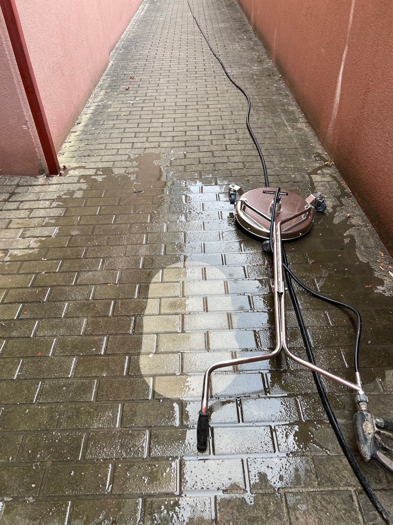Mosiężny czyścik do powierzchni płaskich pod ciśnieniem, z widocznymi śladami wody, na mokrej kostce brukowej w wąskim przejściu między budynkami.