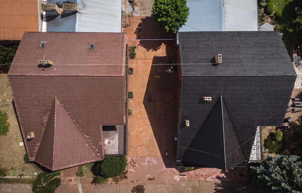 Widok z góry na dwa domy z dachami: jeden z brązową dachówką, drugi z czarną dachówką, oddzielone brukowanym podjazdem z cieniem drzewa.