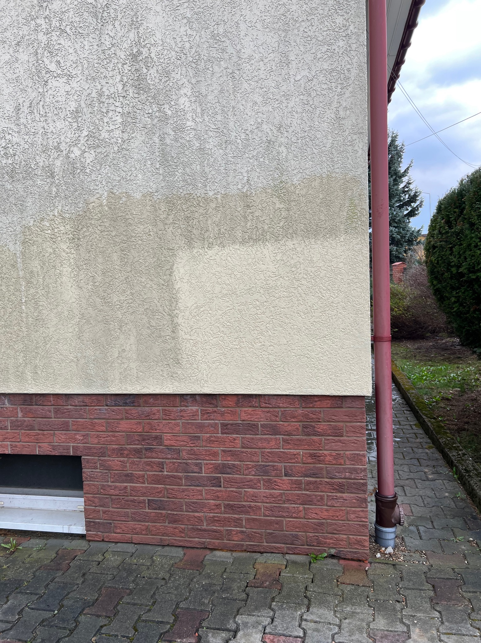 Fragment elewacji budynku z widoczną różnicą w czystości tynku po częściowym umyciu, z czerwonym ceglanym cokołem i rynną.