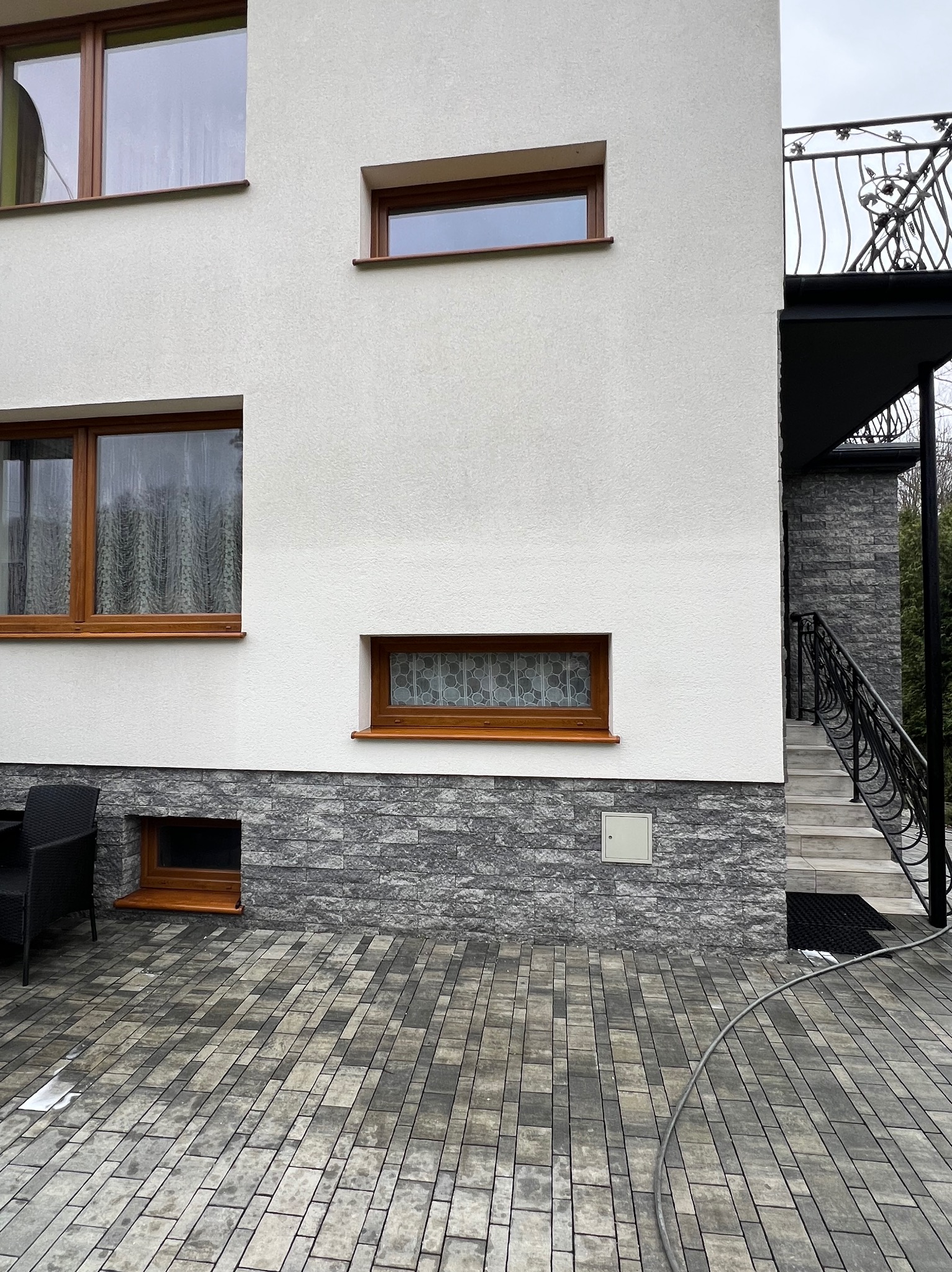 Dwupiętrowy dom z białą elewacją i szarym kamieniem na dole, widoczne okna z drewnianymi ramami, balkon z kutą balustradą, bruk na tarasie i wąż ogrodowy leżący na ziemi.