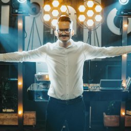 Dj Piter Pan Beatbox - Uśmiechnięty konferansjer z wąsami i okularach, w białej koszuli, stoi na scenie z rozłożonymi rękami, trzymając mikrofon, oświetlony reflektorami.