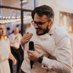 Dj Piter Pan Beatbox - Uśmiechnięty mężczyzna w okularach, ubrany w białą koszulę, trzyma mikrofon i obejmuje drugą osobę podczas wydarzenia z dekoracyjnym oświetleniem w tle.