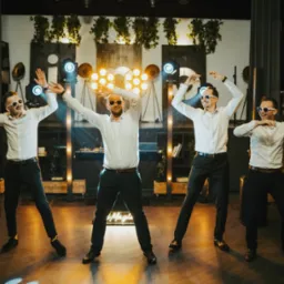 Czterech mężczyzn w białych koszulach i czarnych spodniach, w białych okularach, tańczy na scenie oświetlonej reflektorami i dekoracjami roślinnymi.