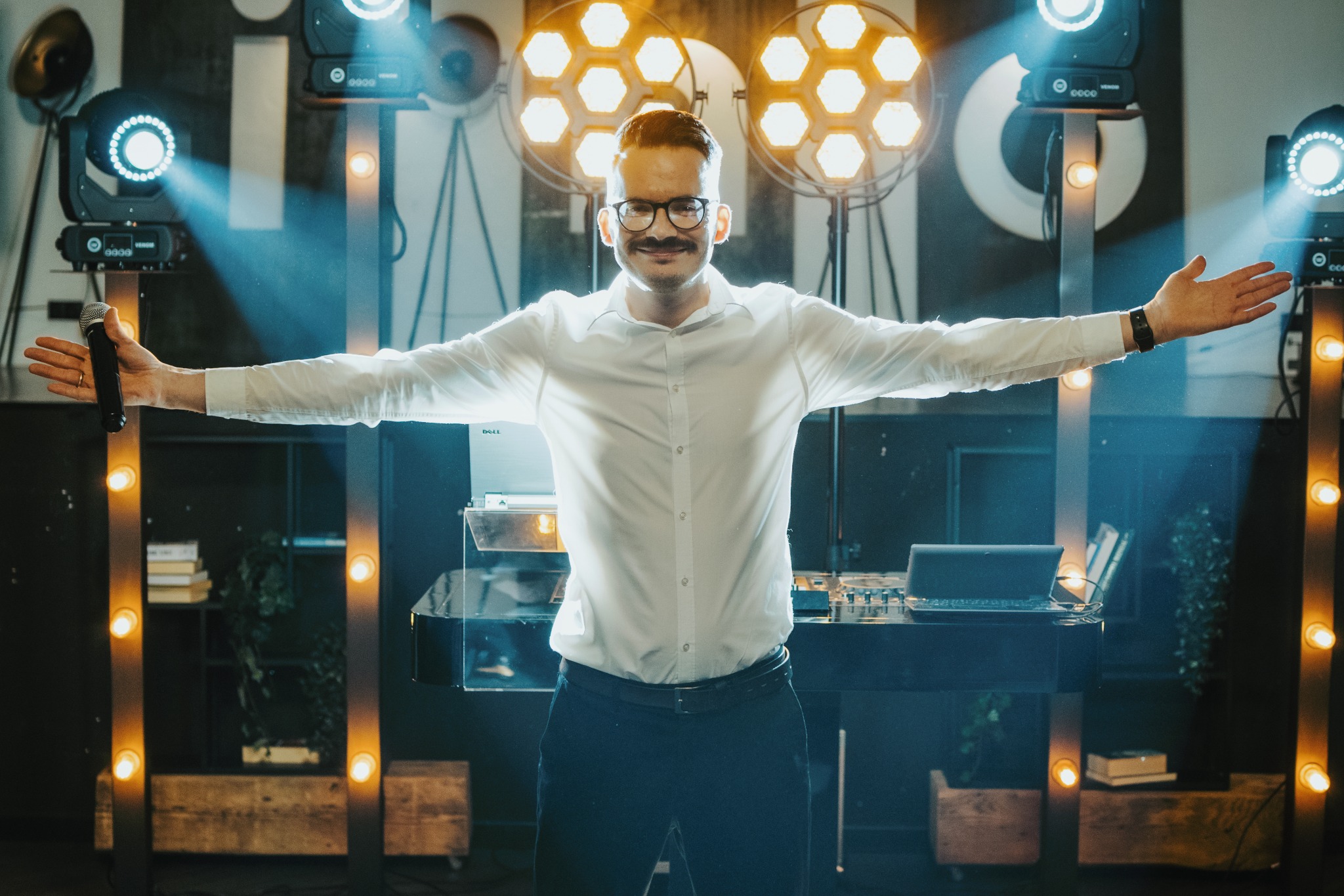 Uśmiechnięty konferansjer z wąsami i okularach, w białej koszuli, stoi na scenie z rozłożonymi rękami, trzymając mikrofon, oświetlony reflektorami.