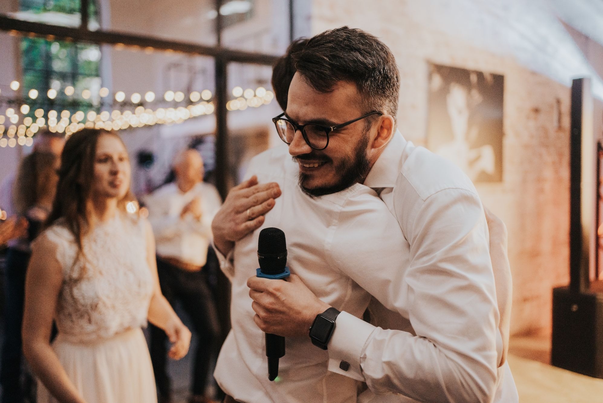 Uśmiechnięty mężczyzna w okularach, ubrany w białą koszulę, trzyma mikrofon i obejmuje drugą osobę podczas wydarzenia z dekoracyjnym oświetleniem w tle.