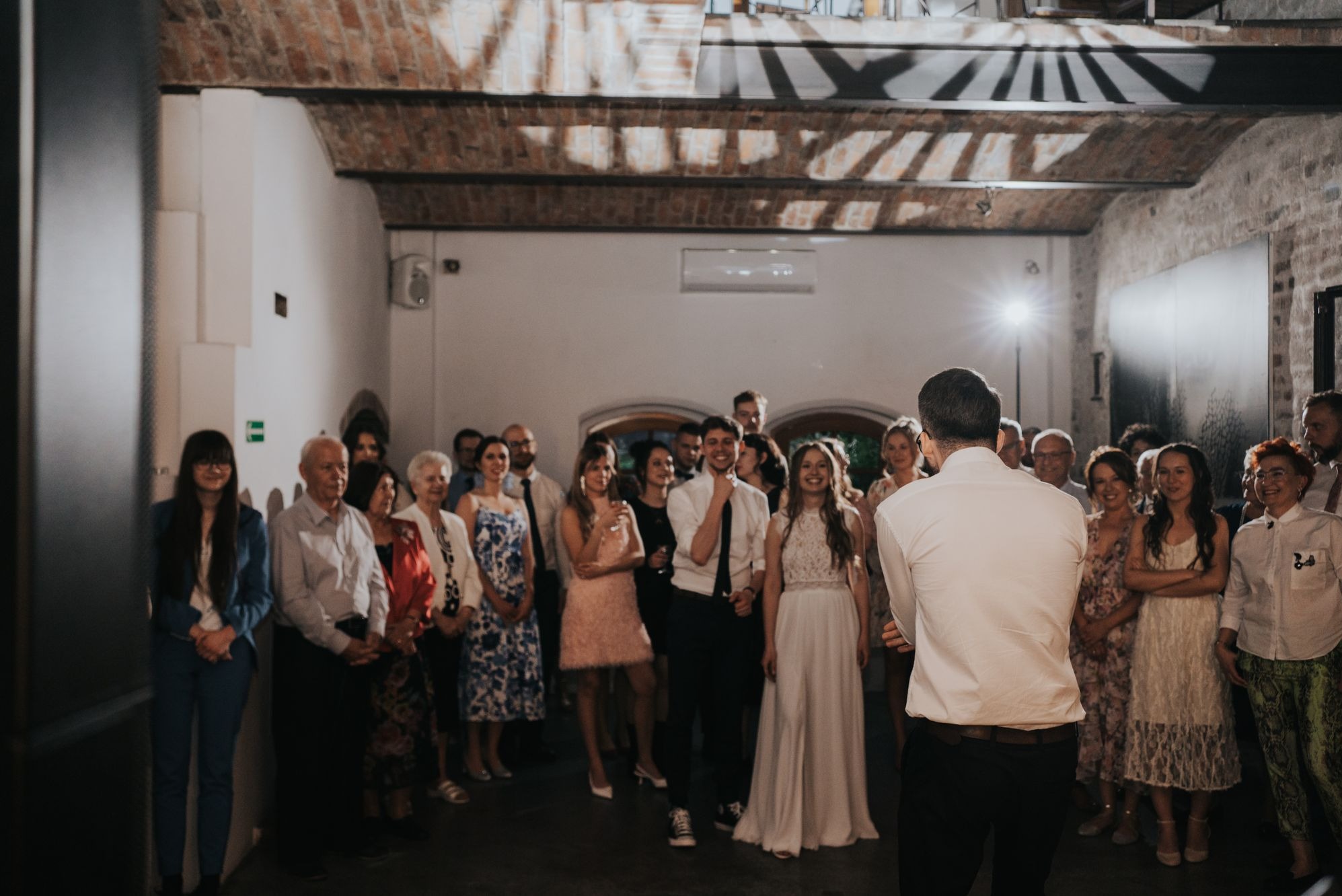 Sala z ceglanym sklepieniem wypełniona gośćmi weselnymi, z prezenterem w białej koszuli stojącym tyłem do obiektywu.