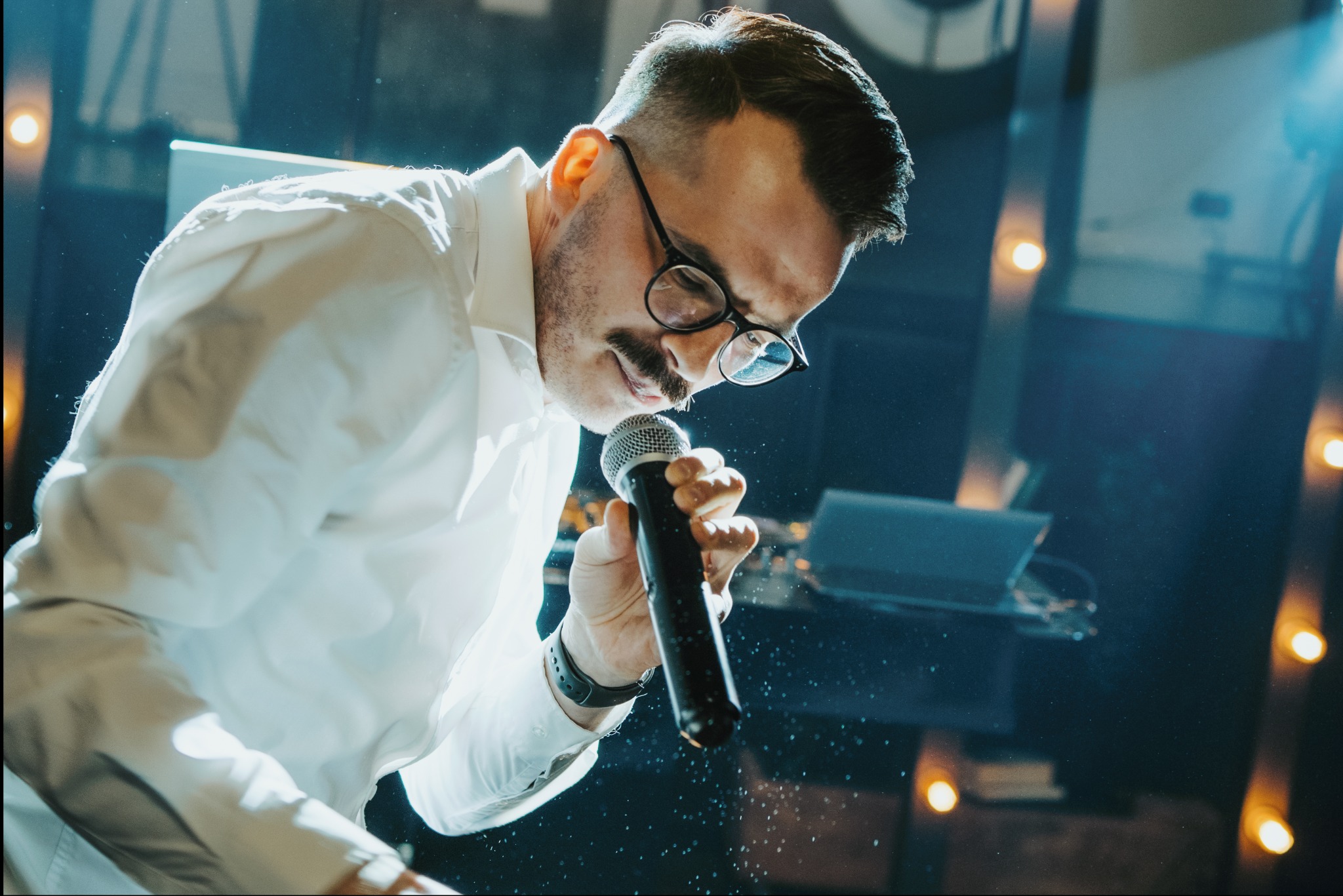 Mężczyzna w okularach i białej koszuli śpiewa do mikrofonu na scenie, oświetlony reflektorami.