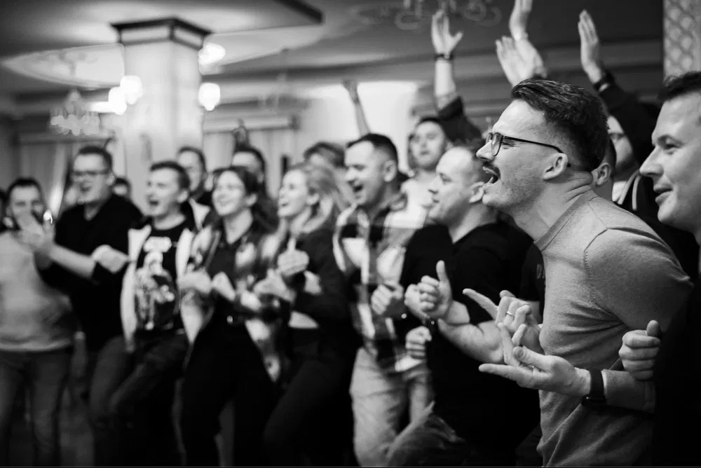 Czarno-białe zdjęcie grupy rozentuzjazmowanych ludzi, prawdopodobnie publiczności, śpiewających lub dopingujących podczas występu, z mężczyzną w okularach i wąsami na pierwszym planie.