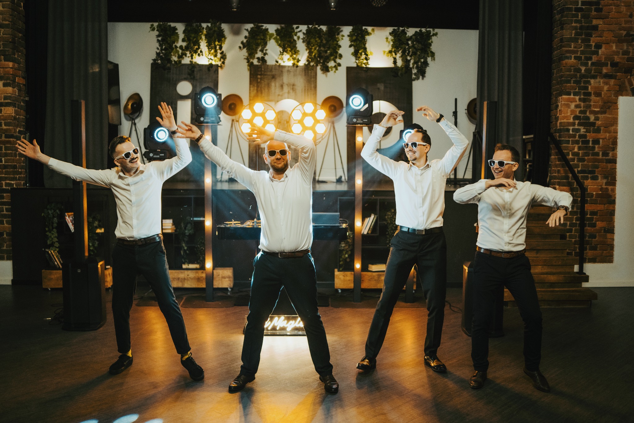 Czterech mężczyzn w białych koszulach i czarnych spodniach, w białych okularach, tańczy na scenie oświetlonej reflektorami i dekoracjami roślinnymi.