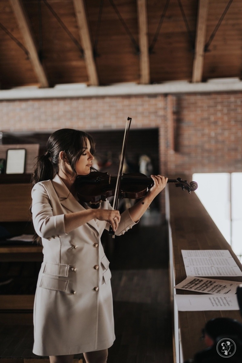 Kobieta w beżowej sukience gra na skrzypcach w pomieszczeniu z ceglaną ścianą i drewnianym sufitem, nuty leżą na stole.