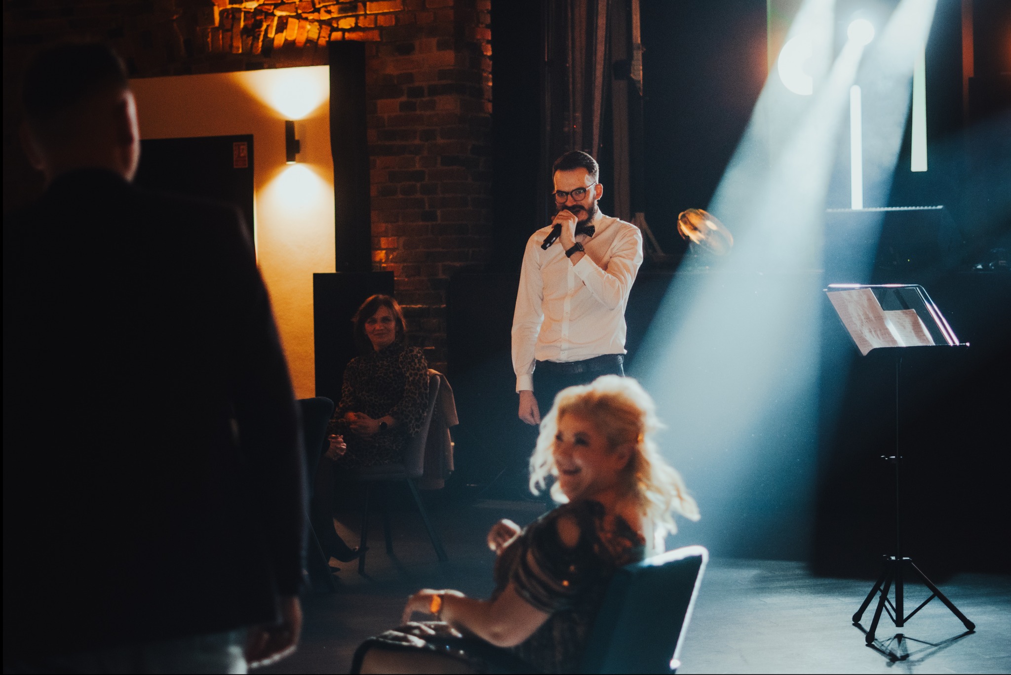 Występ muzyczny w Rybniku: mężczyzna w okularach i muszce śpiewa do mikrofonu na scenie, oświetlony reflektorami, w tle ceglana ściana i publiczność.
