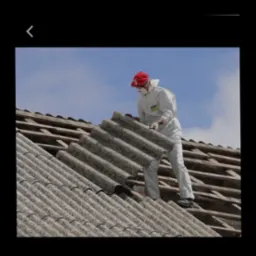 Pracownik w kombinezonie ochronnym i czerwonej czapce demontuje eternit z dachu, widoczne drewniane elementy konstrukcji dachu i błękitne niebo w tle.