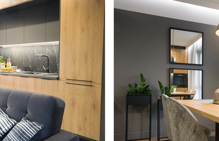 Nowoczesna kuchnia z drewnianymi frontami szafek, czarnym blatem i marmurowym backsplash, widoczny fragment szarej sofy z poduszkami i odbicie kuchni w lustrach na ścianie jadalni.