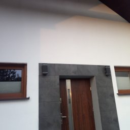 Nik-Bud - Biała elewacja domu z drewnianą stolarką okienną i wejściową, obłożona szarą płytką imitującą kamień wokół drzwi, z widocznymi lampami zewnętrznymi.