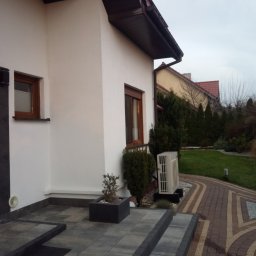 Nik-Bud - Biała elewacja domu jednorodzinnego z brązowymi ramami okiennymi, widoczne schody z szarej kostki brukowej i fragment ogrodu z tujami oraz klimatyzator na zewnątrz budynku.