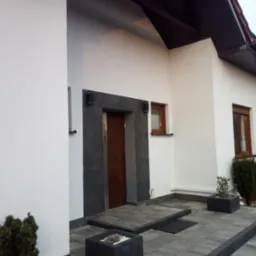 Nowoczesny dom jednorodzinny z jasną elewacją, ciemną stolarką okienną i wejściem wyłożonym szarą kostką brukową, z donicami z roślinami.