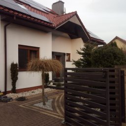 Nik-Bud - Biała elewacja domu jednorodzinnego z brązową stolarką okienną i panelami słonecznymi na dachu, widoczna zza ozdobnej bramy i wybrukowanego podjazdu z elementami dekoracyjnymi.