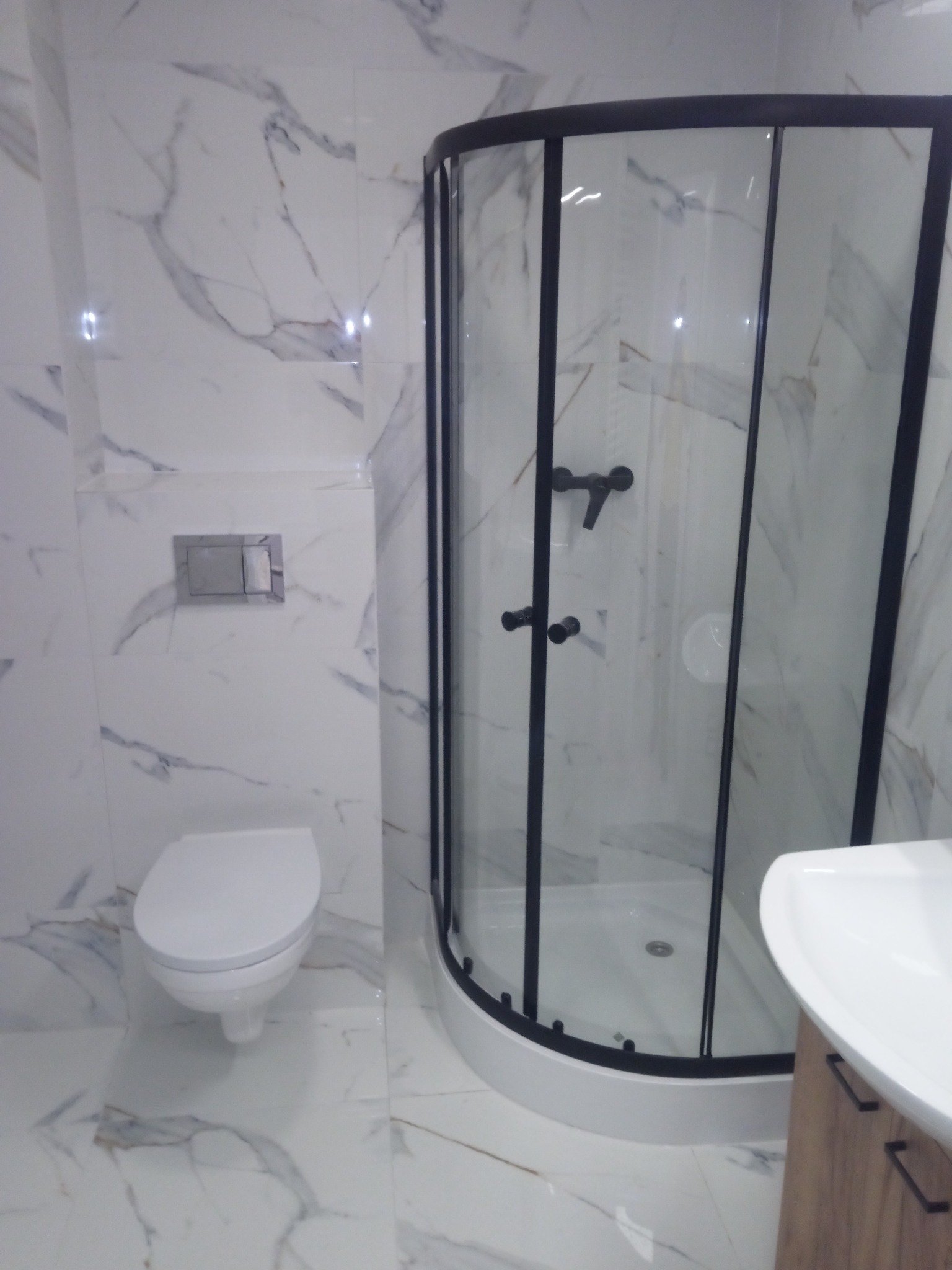 Nowoczesna łazienka z kabiną prysznicową z czarnymi ramami, wiszącą toaletą i umywalką na szafce, wykończona białymi płytkami imitującymi marmur.