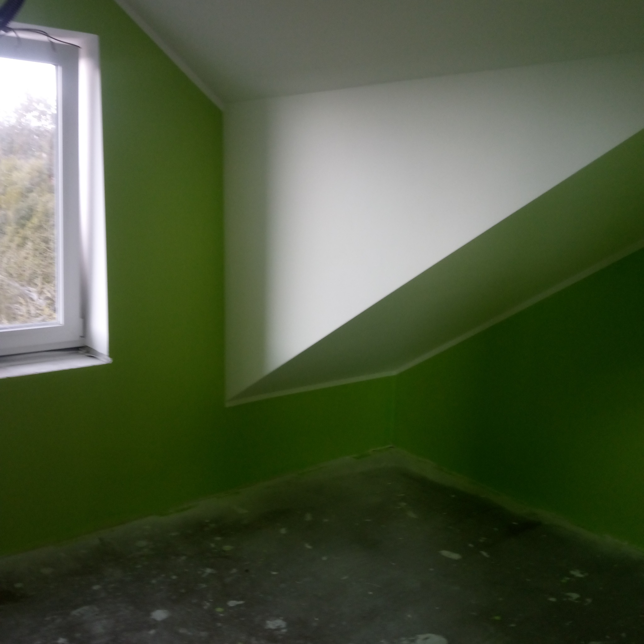 Świeżo pomalowane na zielono wnętrze pokoju na poddaszu z oknem, biały sufit ze skosem, widoczne ślady farby na podłodze.