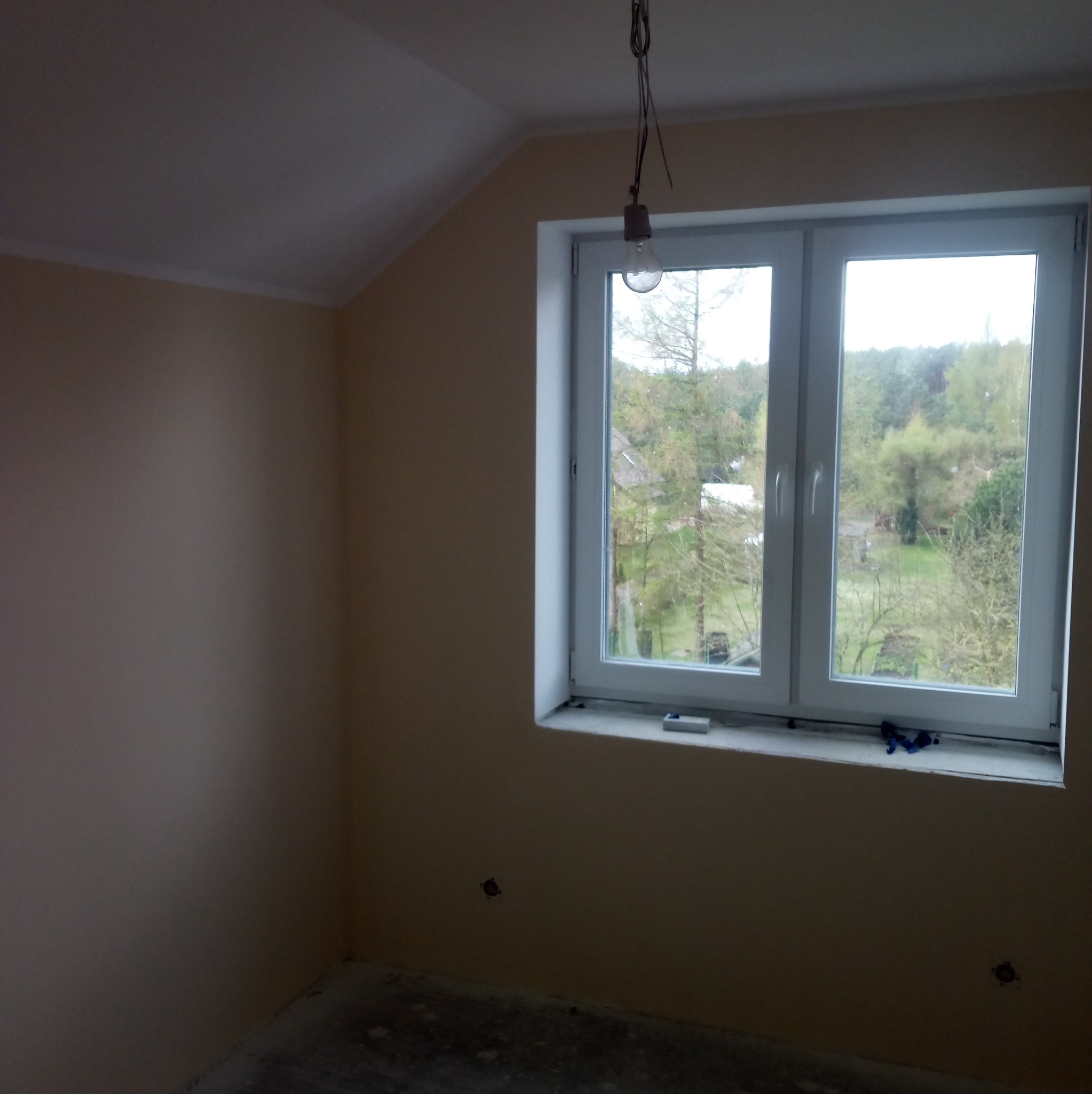 Pokój na poddaszu po malowaniu ścian na beżowo, z białym oknem i żarówką wiszącą na kablu, widok na zewnątrz na zieleń.