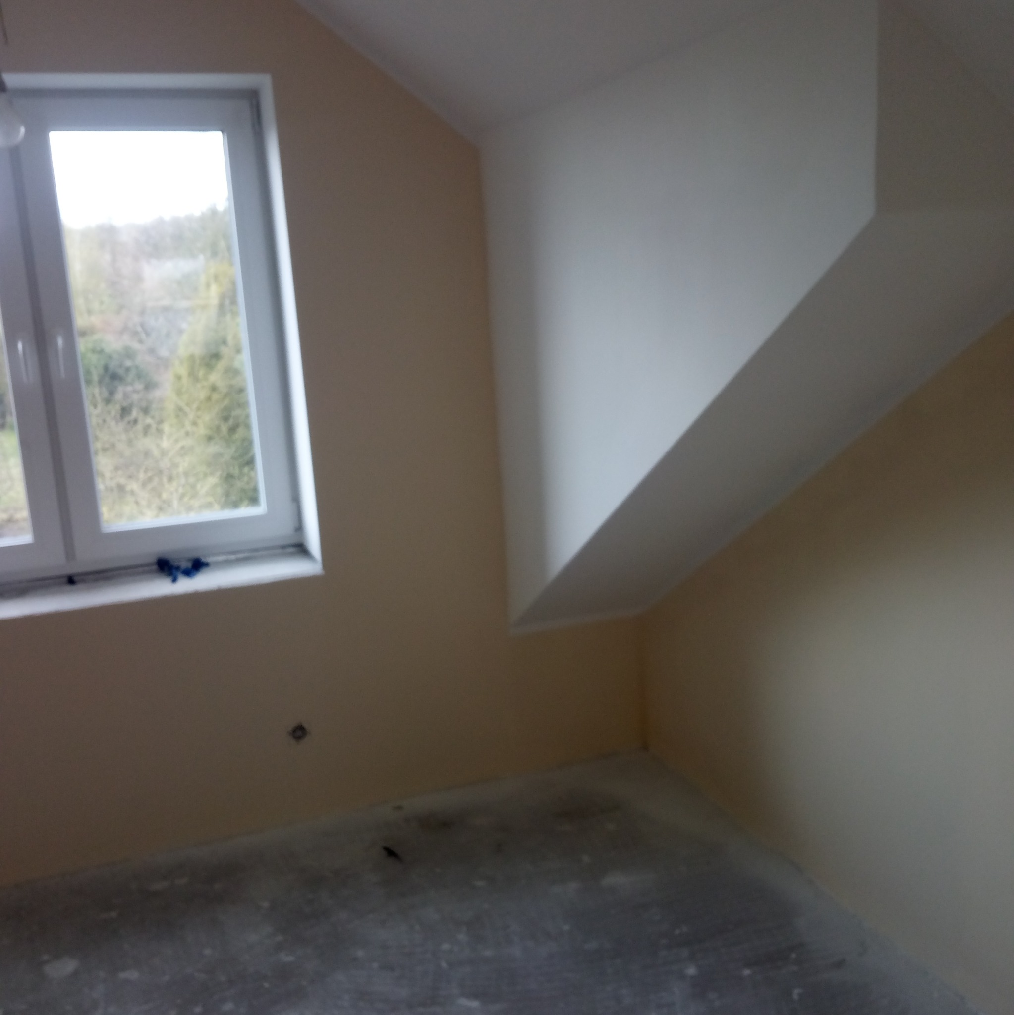 Pomalowane na beżowo poddasze ze skośnym sufitem, widok na okno z widokiem na drzewa, szara, niezakryta podłoga.