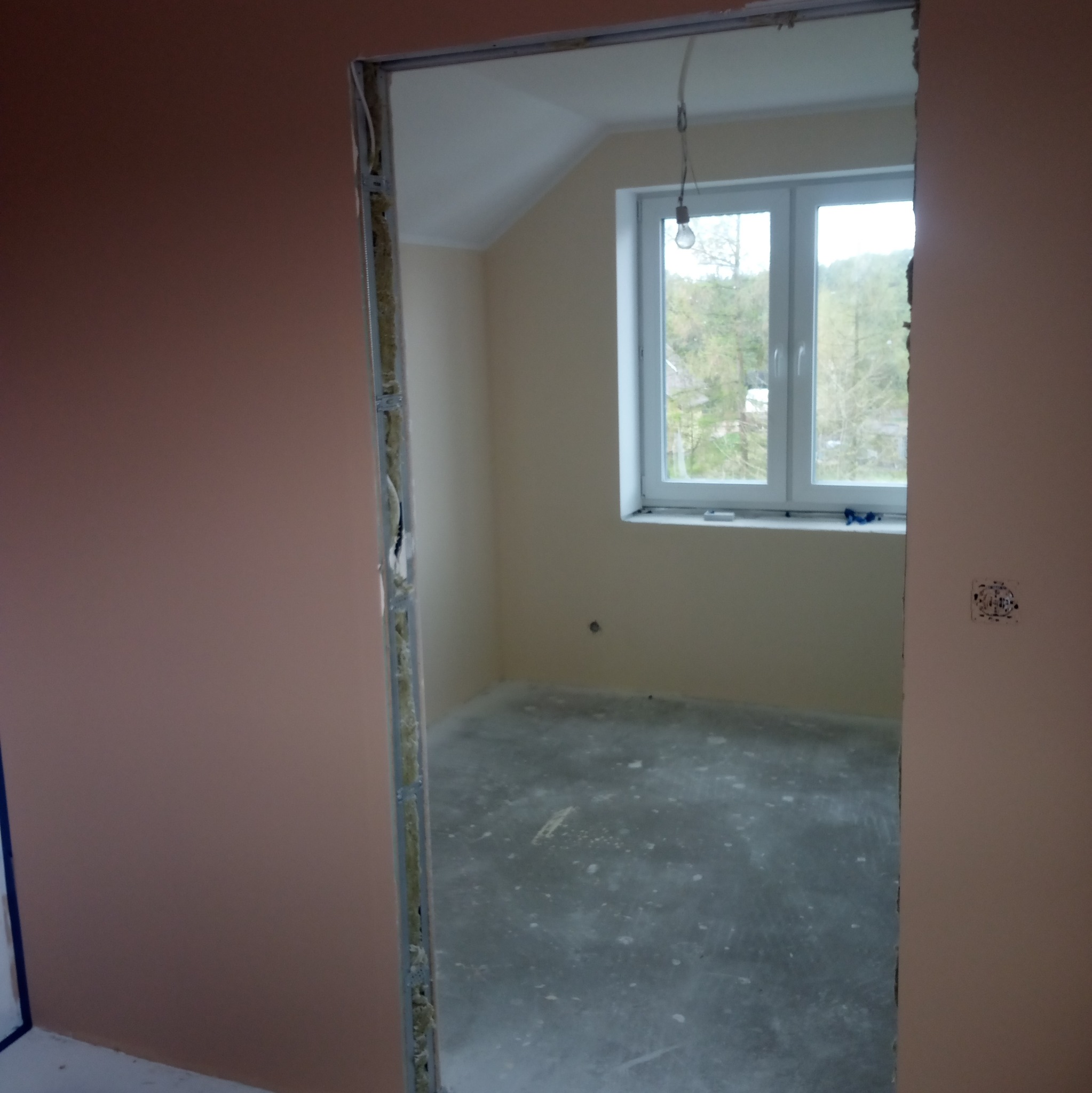 Pokój w trakcie remontu: ściany w odcieniach beżu i różu, widoczne przygotowania do montażu ościeżnicy, surowa wylewka podłogowa, okno z widokiem na zieleń.