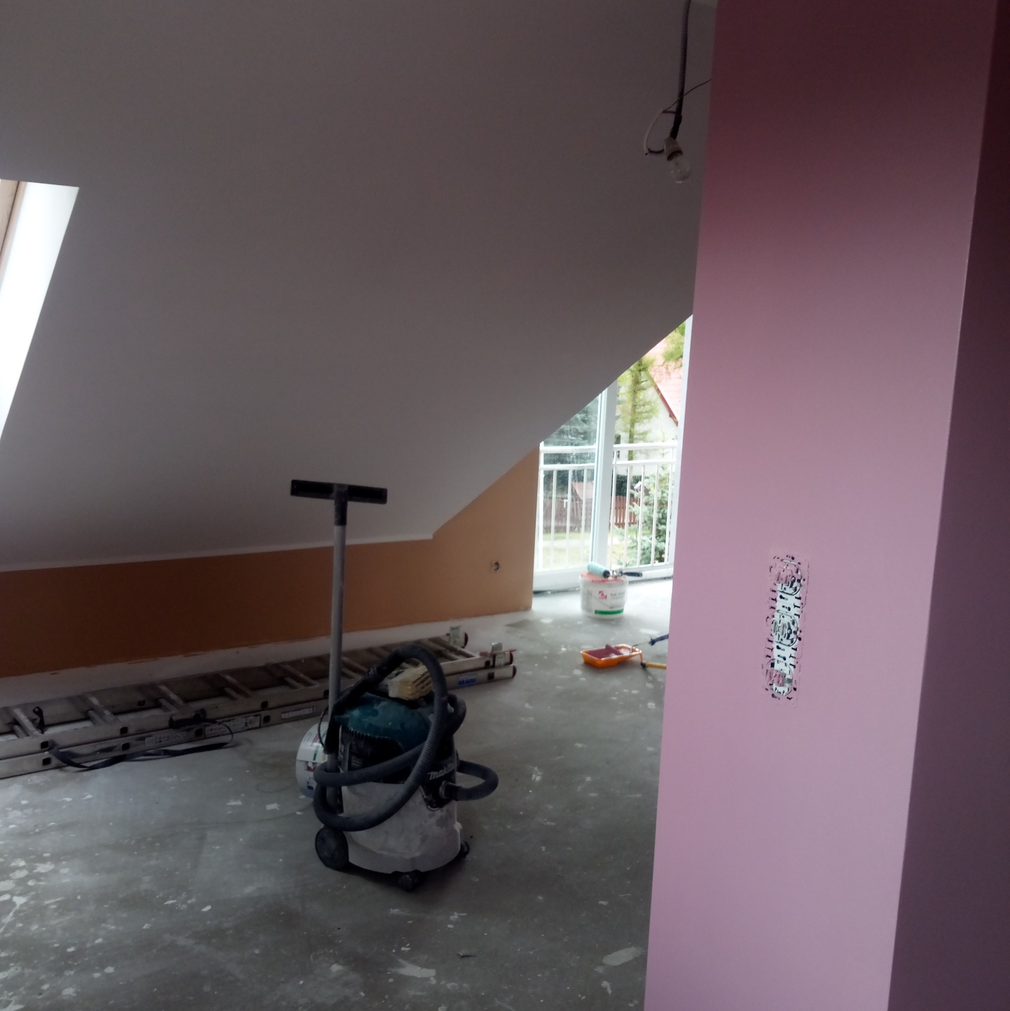 Poddasze w trakcie prac wykończeniowych: widoczna drabina, odkurzacz przemysłowy, ściany częściowo pomalowane na brązowo, a filar na różowo, okno dachowe z widokiem na zieleń.