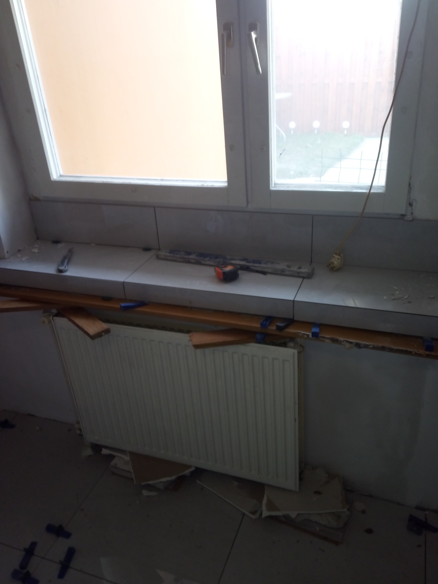 Montaż płytek ceramicznych na parapecie okiennym w trakcie remontu, widoczne listwy dystansowe, grzejnik i narzędzia.