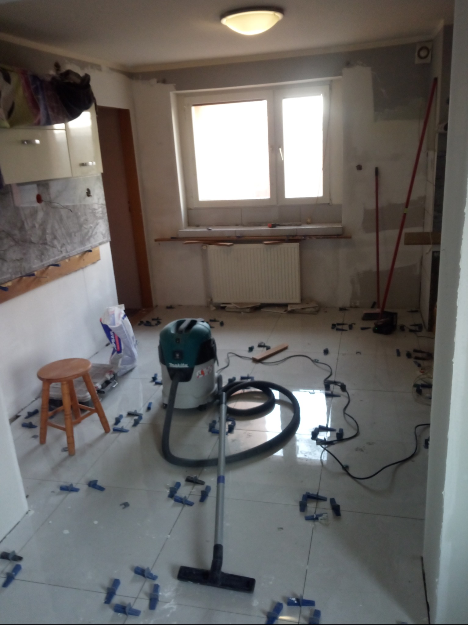 Kuchnia w trakcie remontu: nowe, błyszczące płytki podłogowe z niebieskimi krzyżykami dystansowymi, odkurzacz budowlany Makita i drewniany stołek na tle częściowo wykończonych ścian.