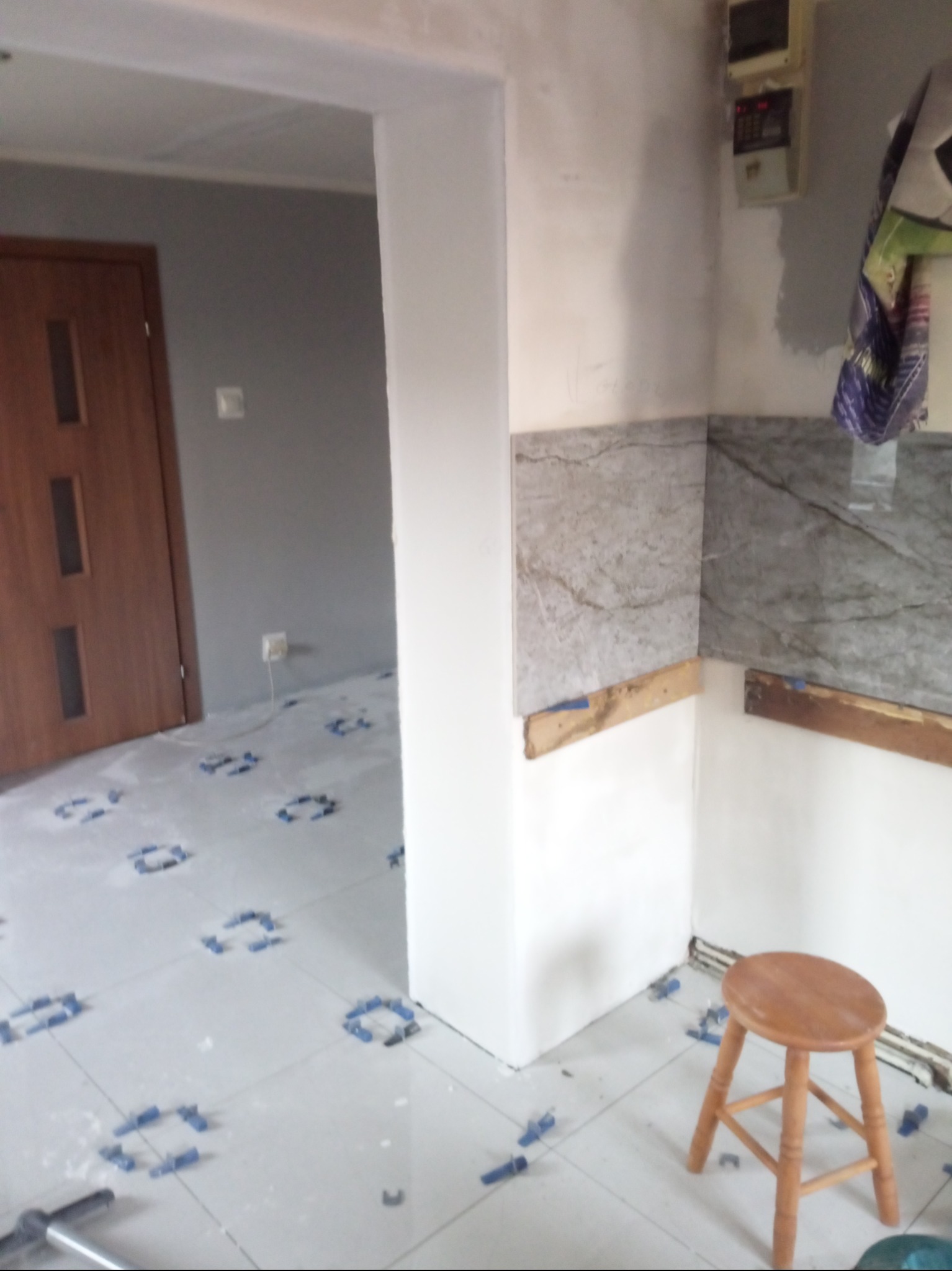 Wnętrze kuchni w trakcie remontu: ułożone płytki podłogowe z niebieskimi krzyżykami dystansowymi, częściowo wykończona ściana z marmurowymi płytkami, drewniany taboret i widoczna instalacja...