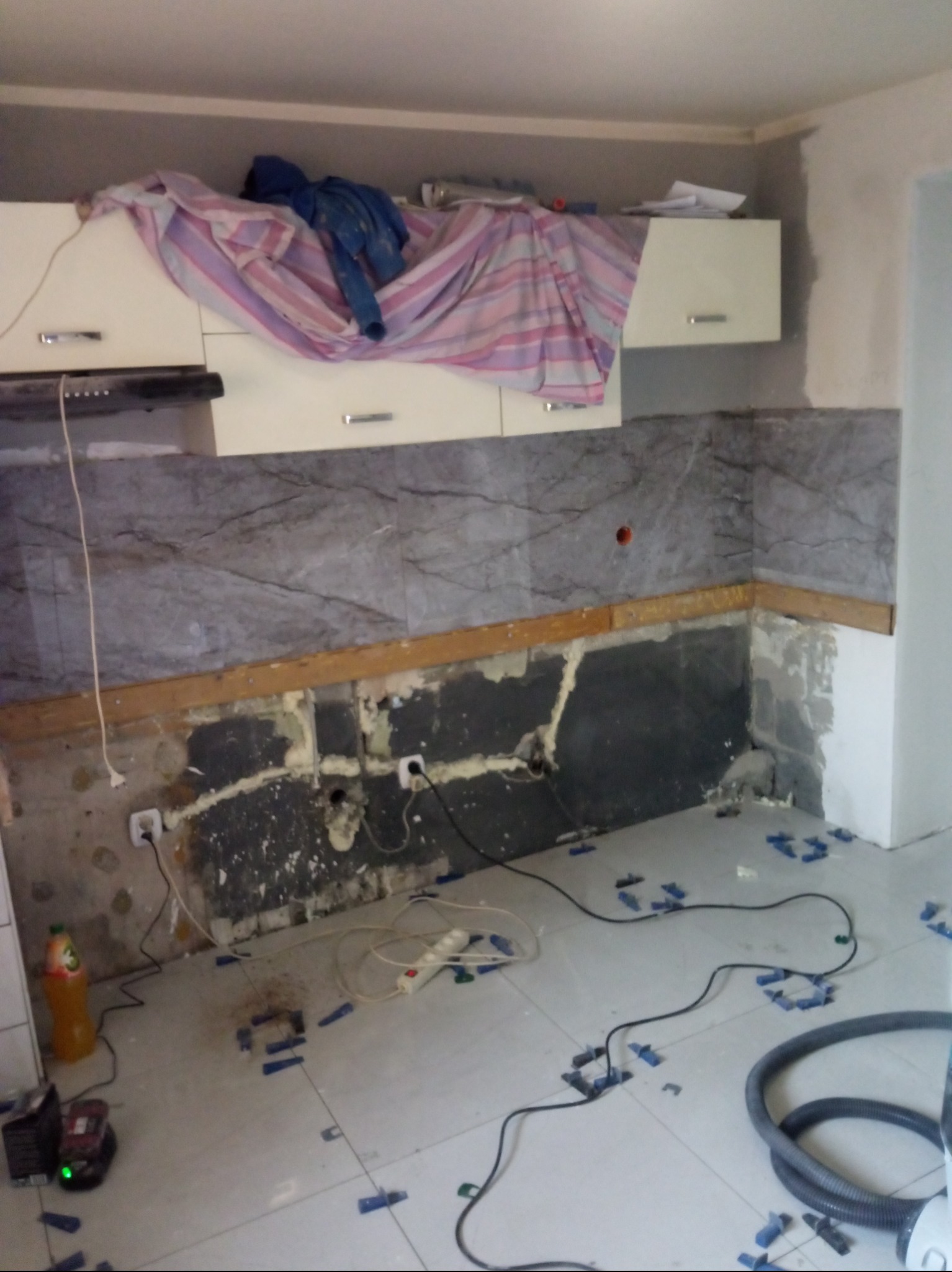 Remont kuchni w trakcie: częściowo ułożone marmurowe płytki na ścianie, widoczne gniazdka elektryczne, listwy montażowe, butelka soku i niebieskie elementy dystansowe na podłodze, ubrania na szafkach.