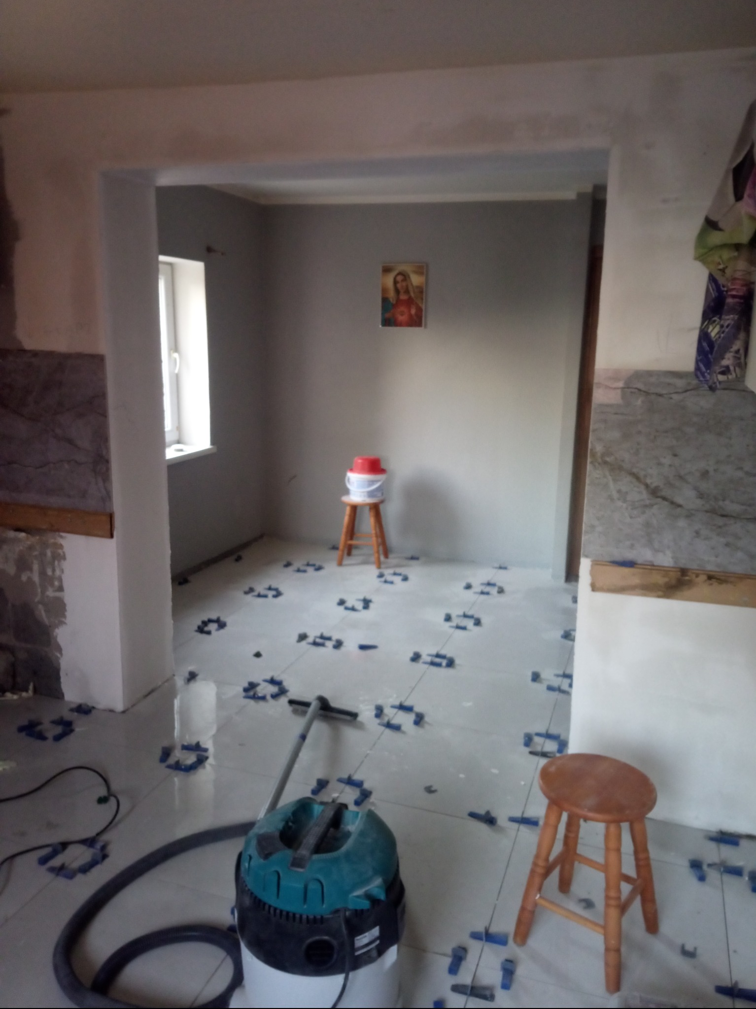 Wnętrze remontowanej kuchni z widocznymi nowymi, błyszczącymi płytkami podłogowymi, niebieskie krzyżyki dystansowe, odkurzacz przemysłowy i taboret.