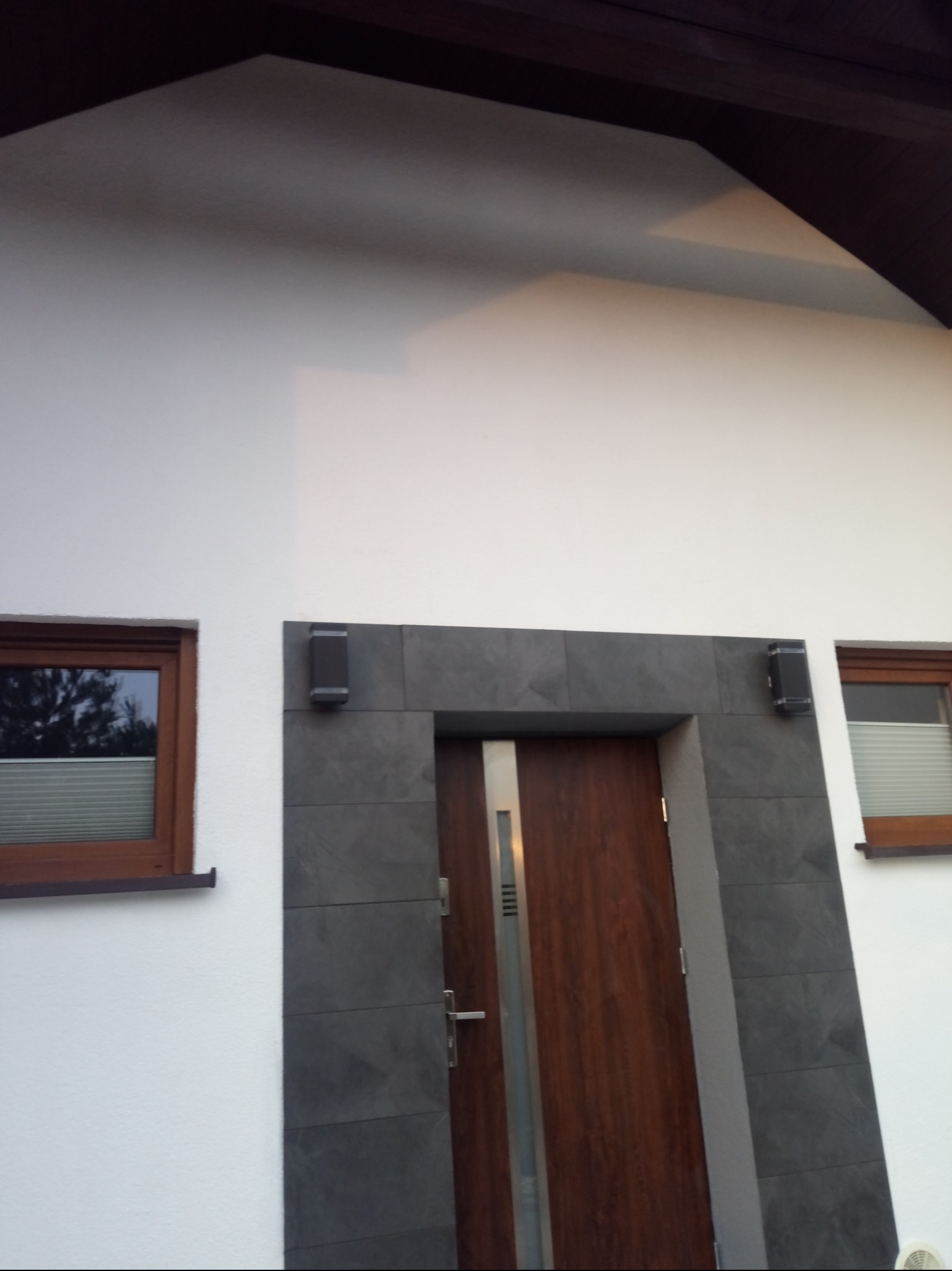 Biała elewacja domu z drewnianą stolarką okienną i wejściową, obłożona szarą płytką imitującą kamień wokół drzwi, z widocznymi lampami zewnętrznymi.