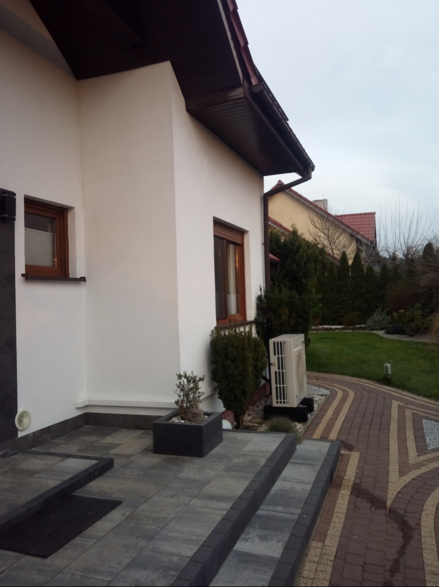 Biała elewacja domu jednorodzinnego z brązowymi ramami okiennymi, widoczne schody z szarej kostki brukowej i fragment ogrodu z tujami oraz klimatyzator na zewnątrz budynku.